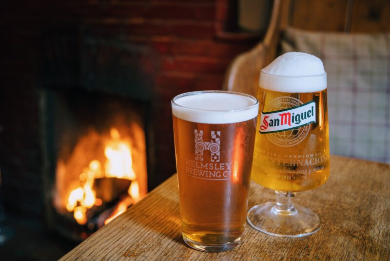 two pints of beer on table bathed in the warm light of a roaring log fire